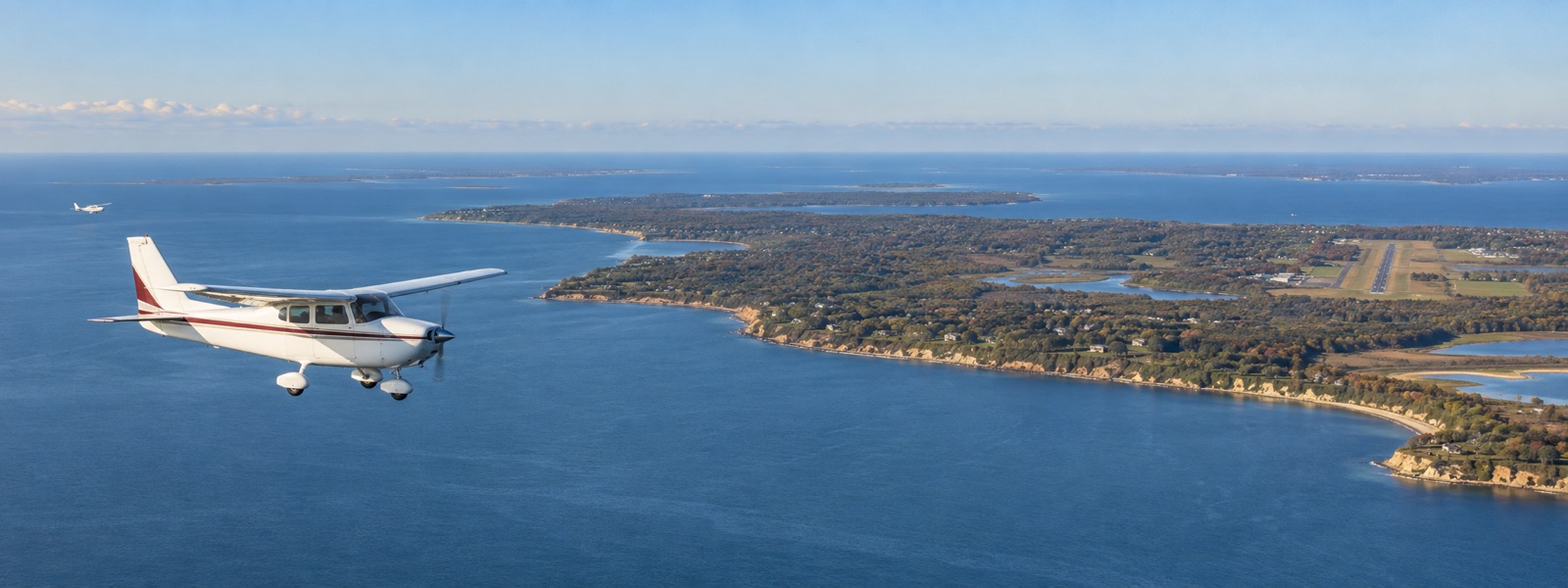 A small high-wing airplane flying over the water toward Martha's Vineyard, with another airplane and the island airport in the distance